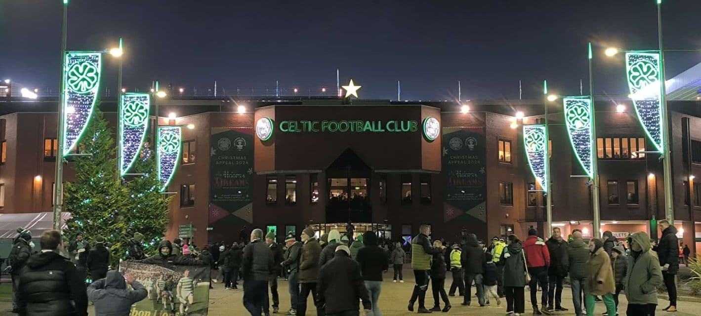 Persone riunite fuori dall'ingresso illuminato dello stadio del Celtic Football Club decorato con luci festive di notte.