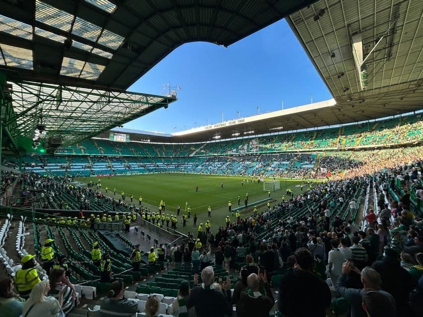 Gli spettatori riempiono le tribune verdi di un grande stadio di calcio durante una partita sotto un cielo azzurro e limpido.
