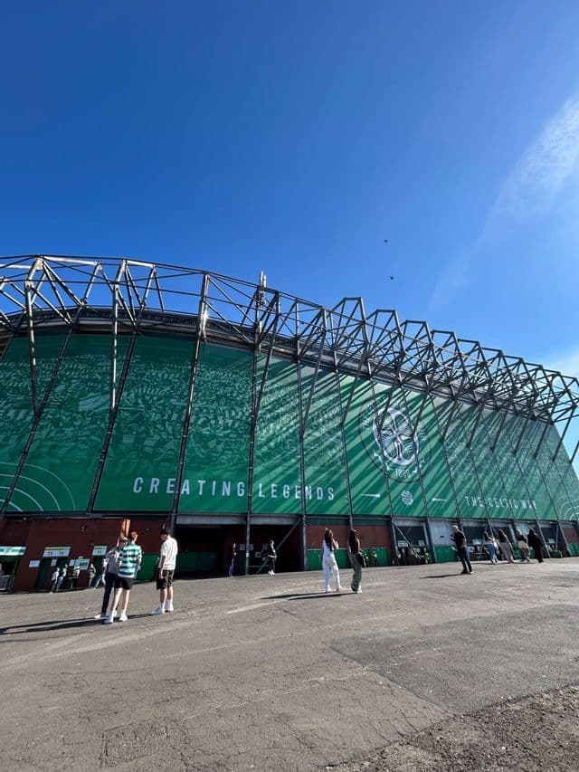 Persone che camminano all'aperto all'esterno di un grande stadio di calcio verde sotto un cielo azzurro e limpido in una giornata di sole.