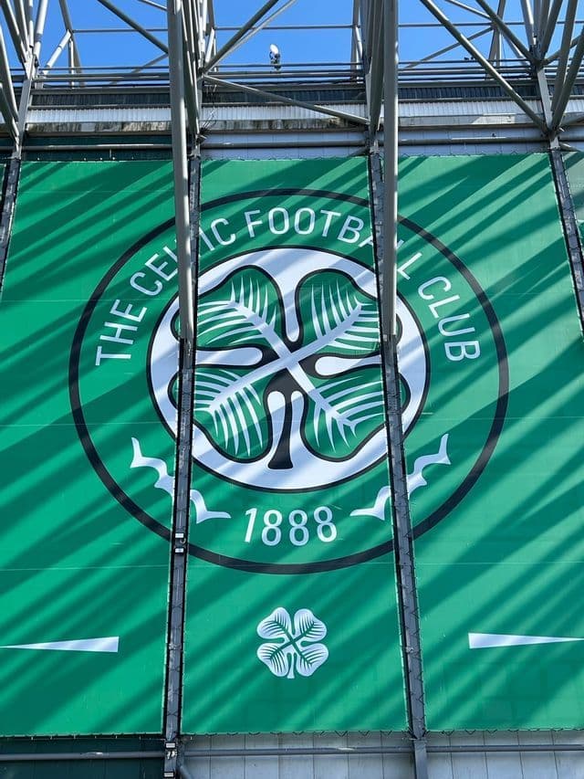 Grande striscione verde con lo stemma del Celtic Football Club montato sulla facciata di uno stadio con travi strutturali in metallo.
