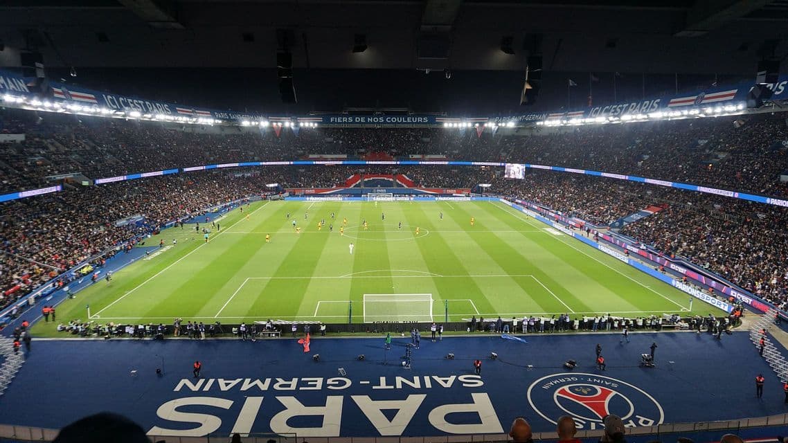 Giocatori di calcio in competizione su un campo verde all'interno di uno stadio affollato sotto forti riflettori di notte.