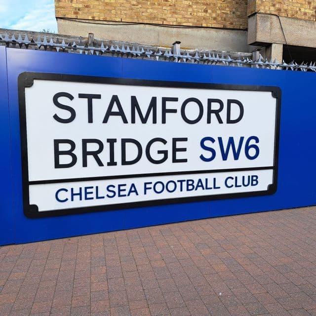 Un cartello stradale con la scritta Stamford Bridge SW6 Chelsea Football Club montato su un muro blu lungo un percorso lastricato.