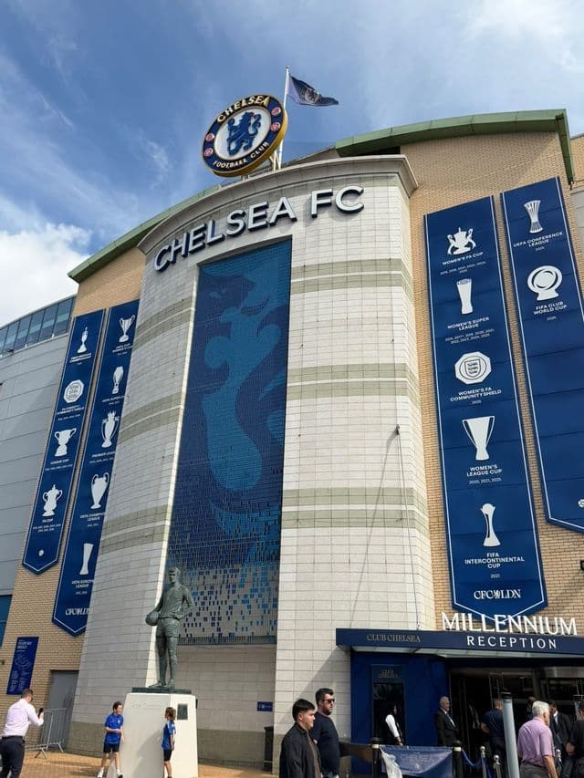Facciata dello stadio di calcio con grandi striscioni di trofei blu, uno stemma della squadra e una statua di bronzo all'ingresso.