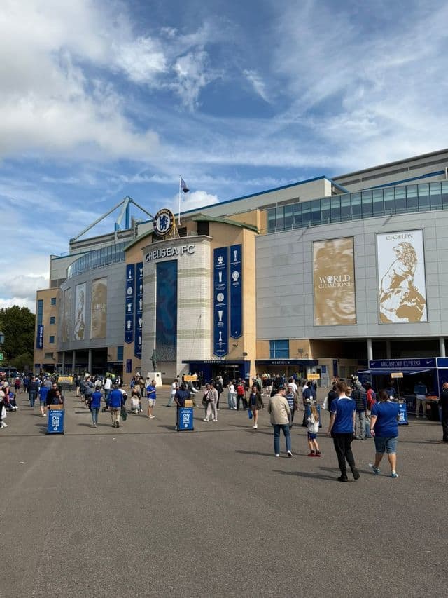 Folla di persone che cammina in una piazza aperta fuori da un moderno stadio di calcio con grandi striscioni in una giornata di sole