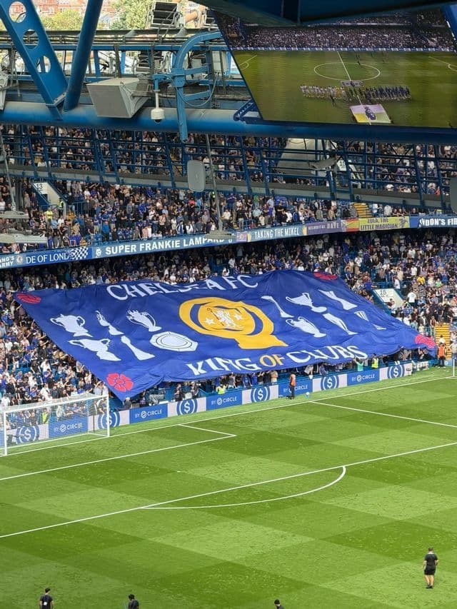 I tifosi di calcio sugli spalti dello stadio tengono in alto un grande striscione blu che mostra trofei dietro la porta del campo.