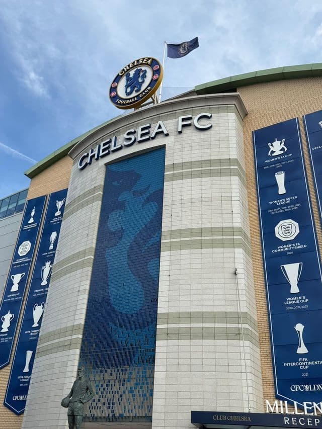 L'esterno di uno stadio di calcio con insegna del Chelsea FC, grandi striscioni di trofei blu e una statua di bronzo di un giocatore.