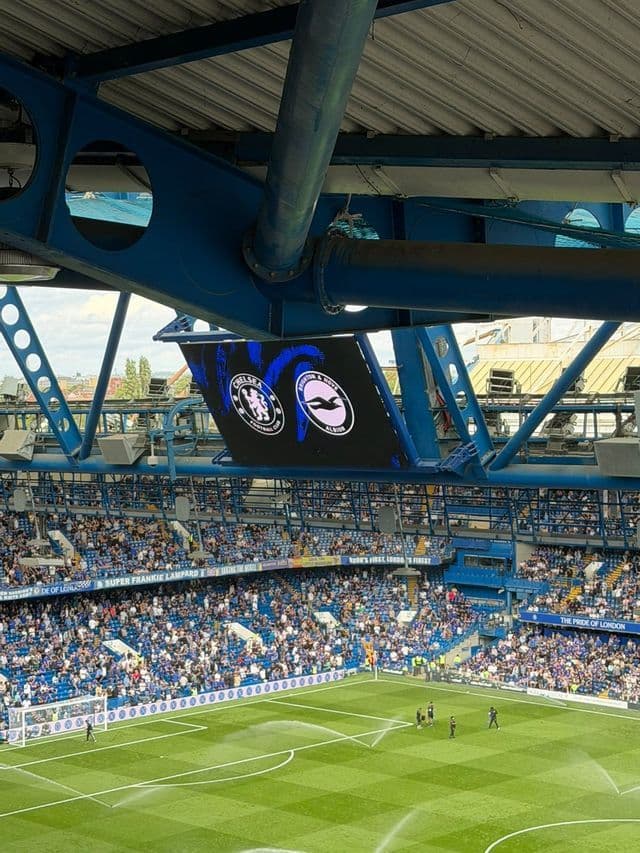 Una vista dagli spalti superiori di uno stadio di calcio affollato che si affaccia sul campo con un grande tabellone segnapunti.