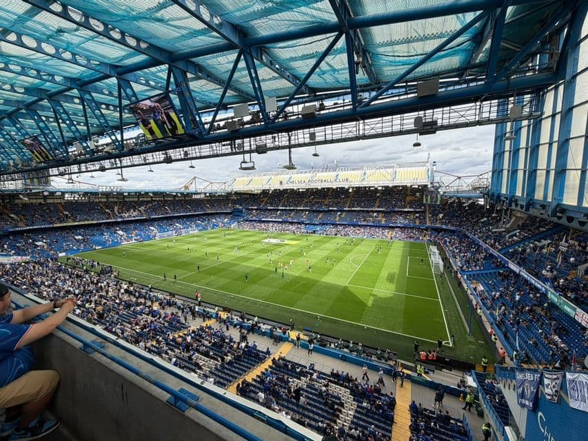 Spettatori sugli spalti blu dello stadio guardano i giocatori su un campo da calcio verde da una vista rialzata sotto un tetto blu.