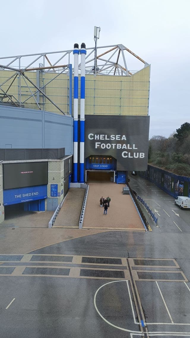 L'esterno di uno stadio di calcio con grandi insegne, tunnel d'ingresso e persone che camminano lungo una passerella rialzata.