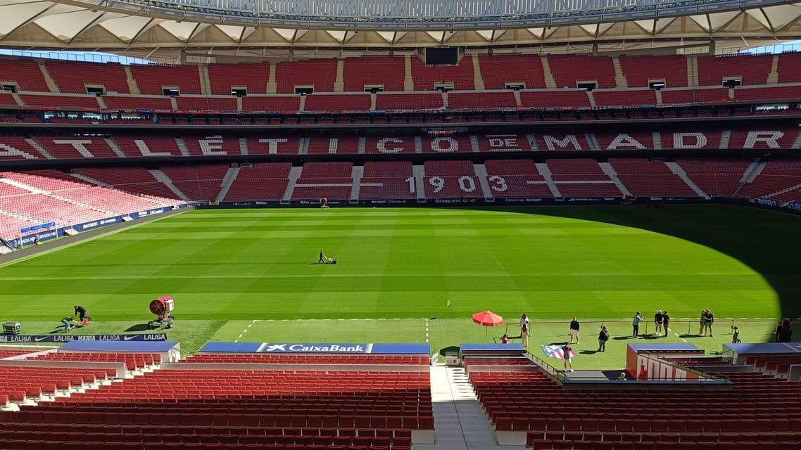 Uno stadio sportivo vuoto con gradinate rosse attorno a un campo verde vibrante in manutenzione in una giornata di sole