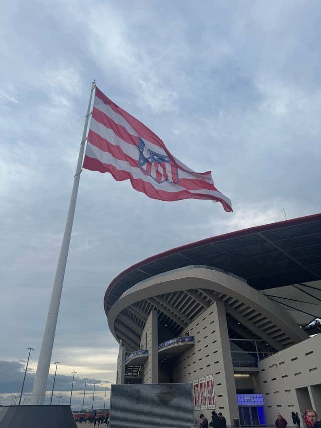 Una bandiera a strisce bianche e rosse sventola su un alto palo fuori da uno stadio moderno contro un cielo coperto.