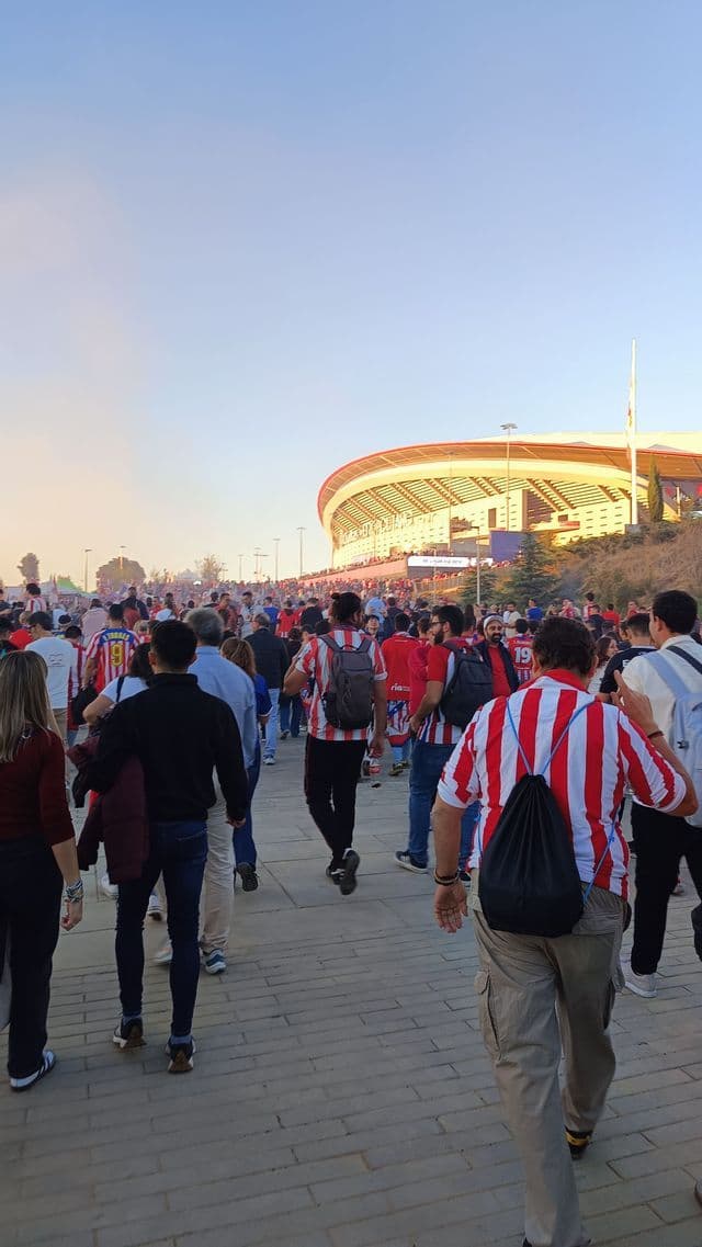 Una folla di tifosi di calcio cammina lungo un ampio viale lastricato verso un moderno stadio sotto un cielo sereno