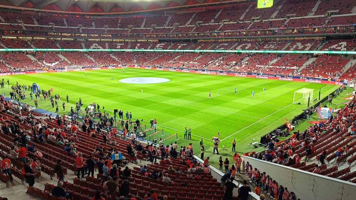 Giocatori di calcio che si allenano su un campo verde brillante all'interno di un grande stadio pieno di spalti rossi.