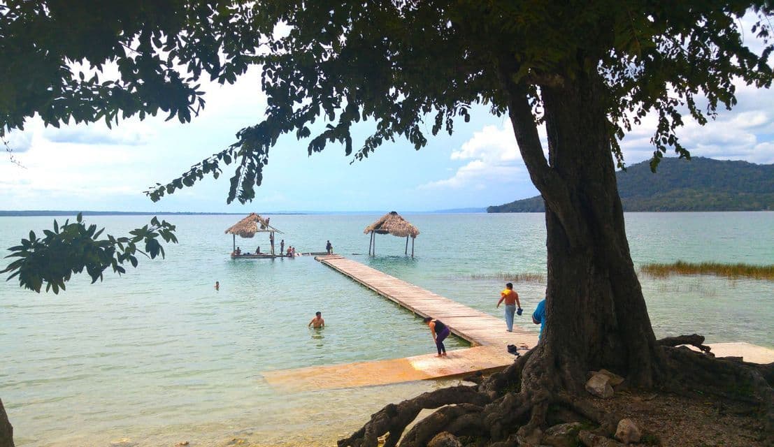Menschen schwimmen und gehen auf einem Holzsteg mit Strohhütten im ruhigen Wasser, eingerahmt von einem großen Seeuferbaum.