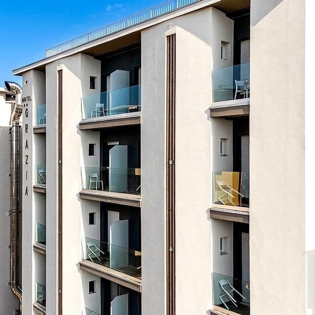 Vista esterna della facciata di un moderno hotel a più piani con balconi dotati di parapetti in vetro e sedie.
