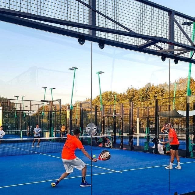 Uomini in abbigliamento sportivo che giocano una partita su un campo da padel all'aperto di colore blu, visti attraverso una parete di vetro.