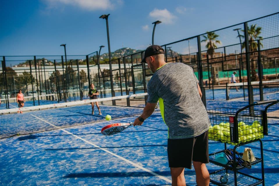 Un uomo gioca a padel con una racchetta su un campo blu all'aperto vicino a un cesto di palline durante una sessione di allenamento.