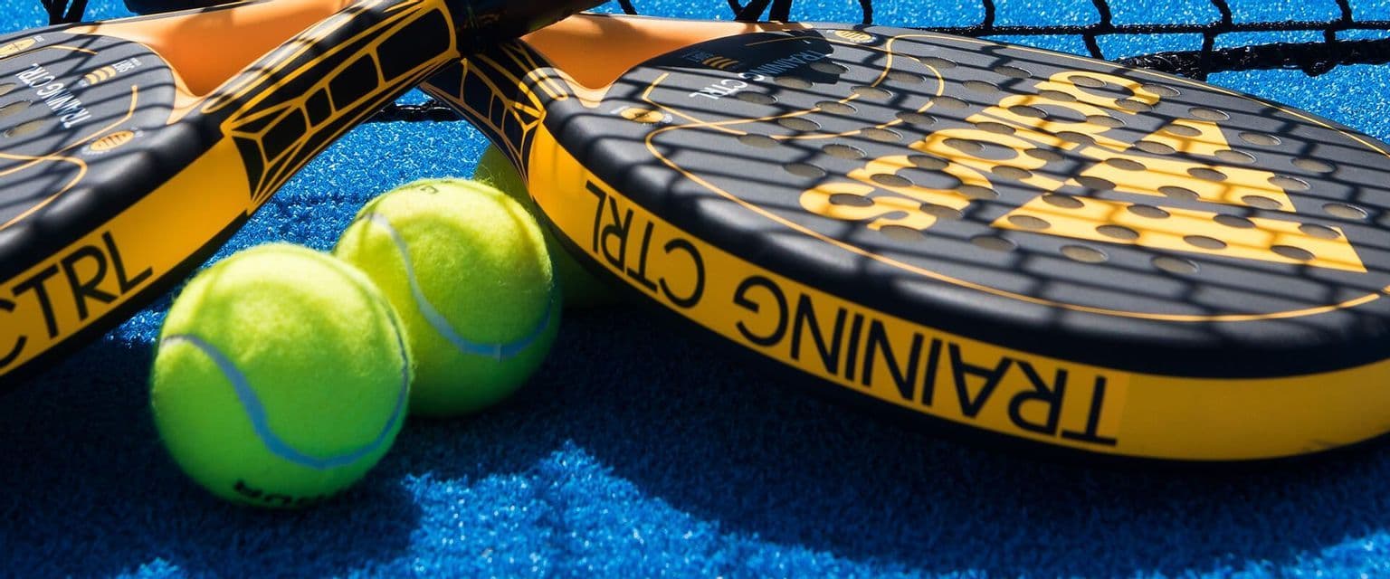 Racchette da padel gialle e nere appoggiate accanto a palline al neon su un campo in erba sintetica blu vicino alla rete