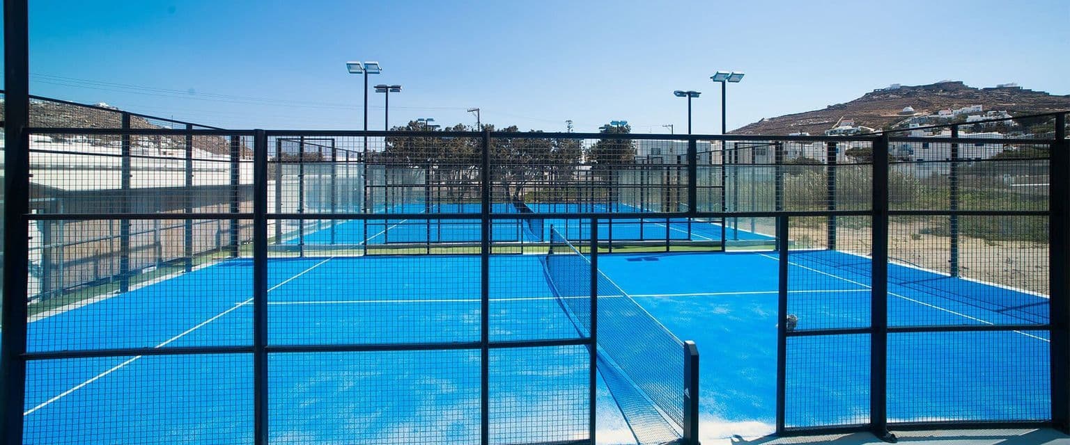 Campi da padel all'aperto di colore blu recintati da una rete metallica nera e pareti di vetro sotto un cielo limpido e luminoso.