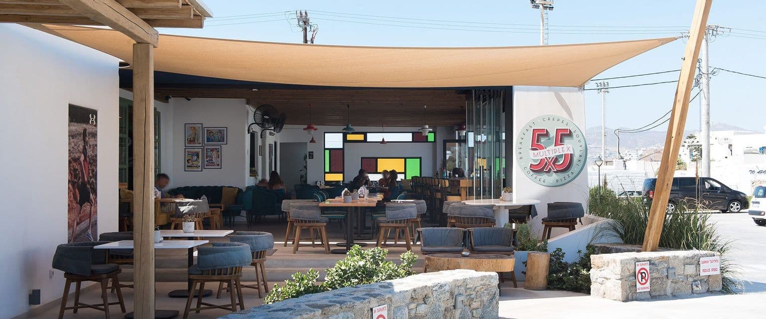 Un patio di un ristorante all'aperto con tavoli in legno, sedie con cuscini e una vela parasole in alto in una giornata di sole
