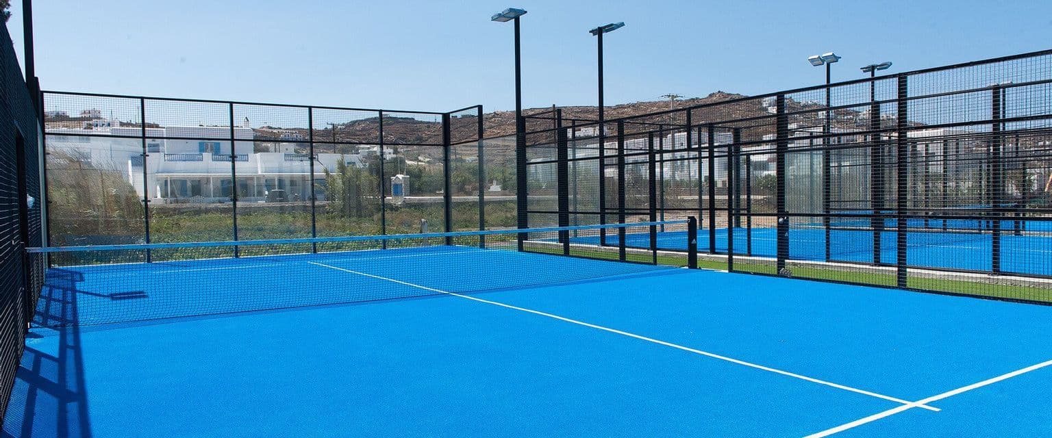 Campi da padel all'aperto vuoti con erba sintetica blu brillante e recinzioni in rete nera sotto un cielo azzurro.