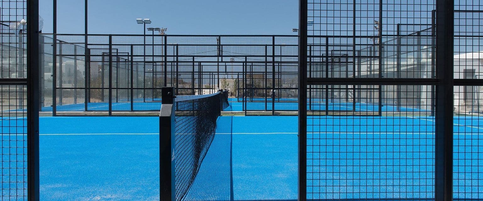 Campi da padel all'aperto vuoti con erba sintetica blu brillante e recinzioni in rete metallica nera sotto un cielo sereno.