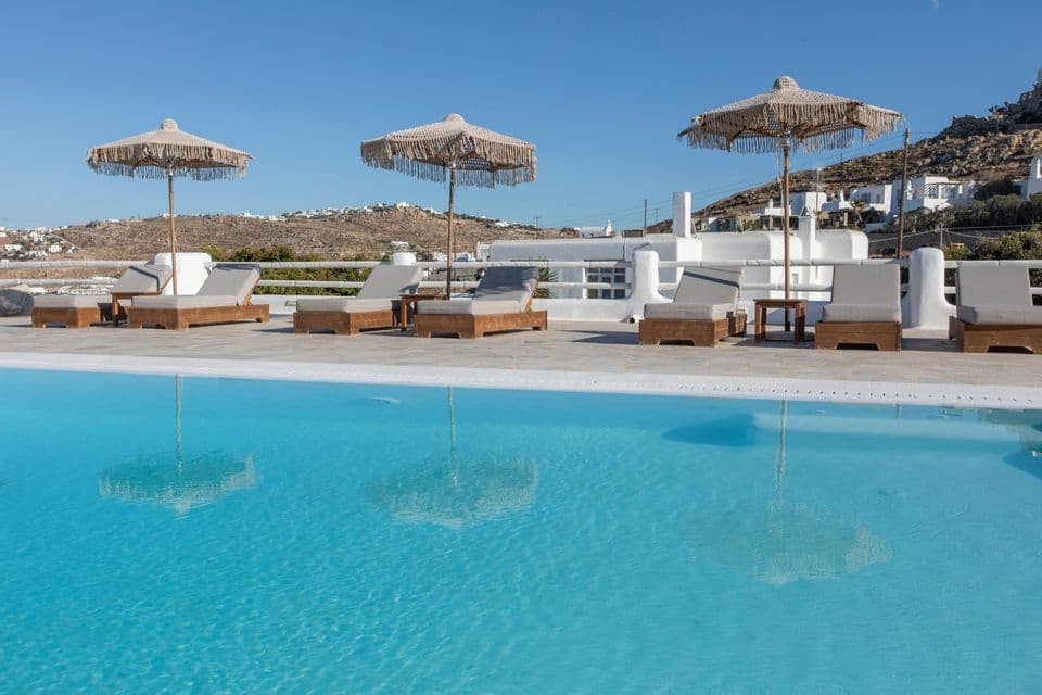 Lettini con ombrelloni con frange disposti vicino a una piscina blu cristallina sotto un cielo azzurro.