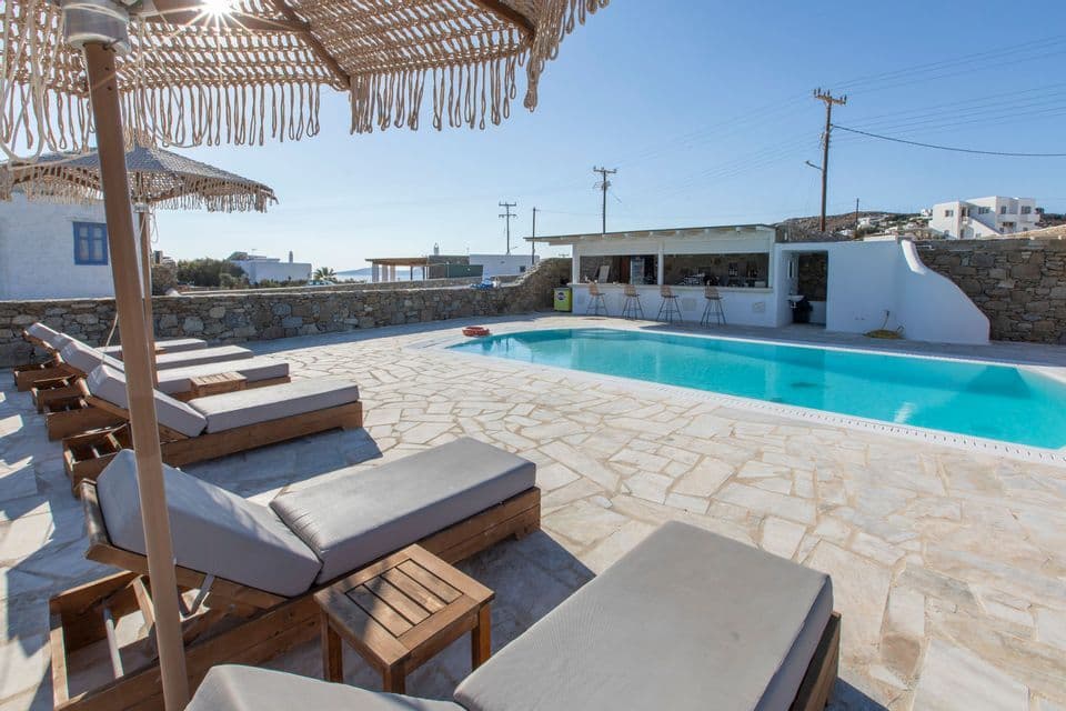 Lettini disposti attorno a una luminosa piscina blu su un patio in pietra con bar a bordo piscina sotto un cielo azzurro e limpido