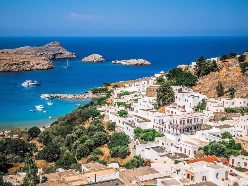 Edifici bianchi sulla collina che si affacciano su una baia dal mare turchese con barche ormeggiate e isole rocciose sotto un cielo azzurro e limpido.