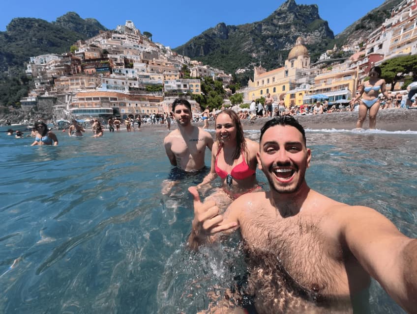 Un viaje en grupo de amigos de WeRoad sonriendo y nadando en el mar cristalino frente a un pueblo costero lleno de gente en la colina.
