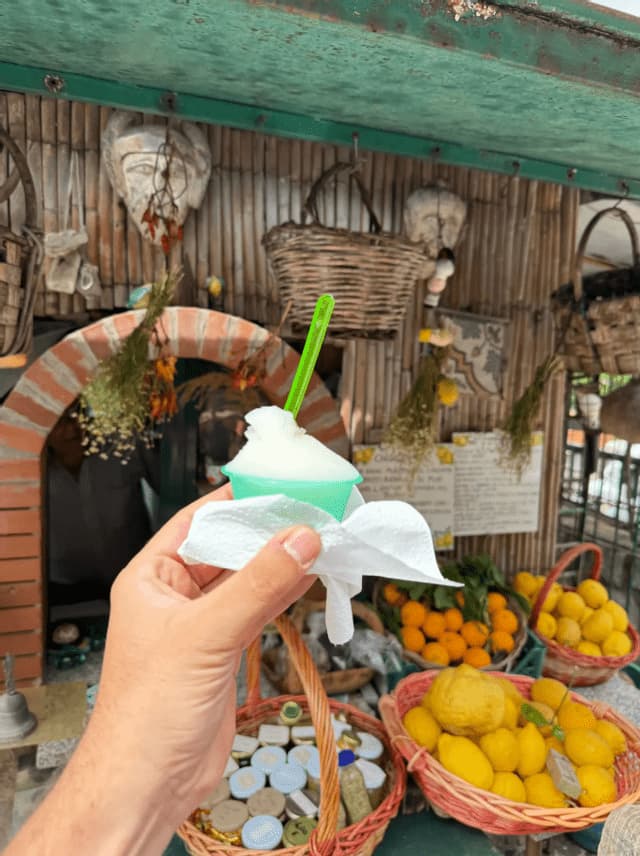 Una mano sostiene una taza de granizada blanca frente a un puesto de fruta rústico con cestas de limones y naranjas frescos