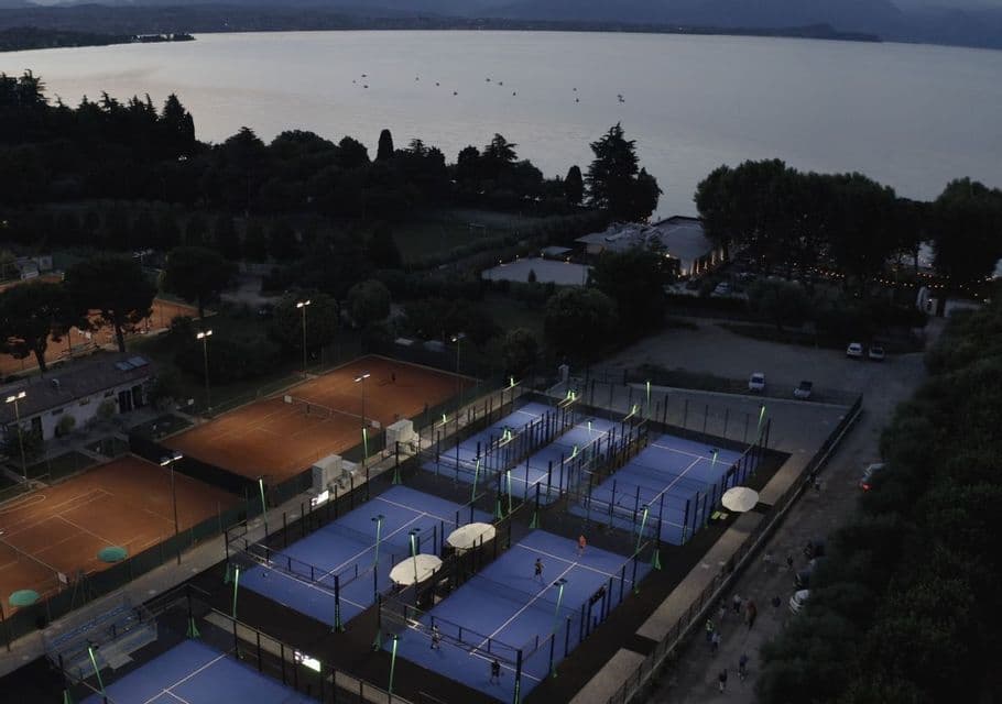 Illuminated blue padel courts and clay tennis courts next to a large lake at dusk.