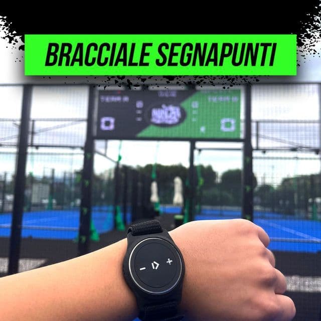 A close-up of a person's wrist wearing a black scorekeeping device in front of outdoor padel courts and a scoreboard.