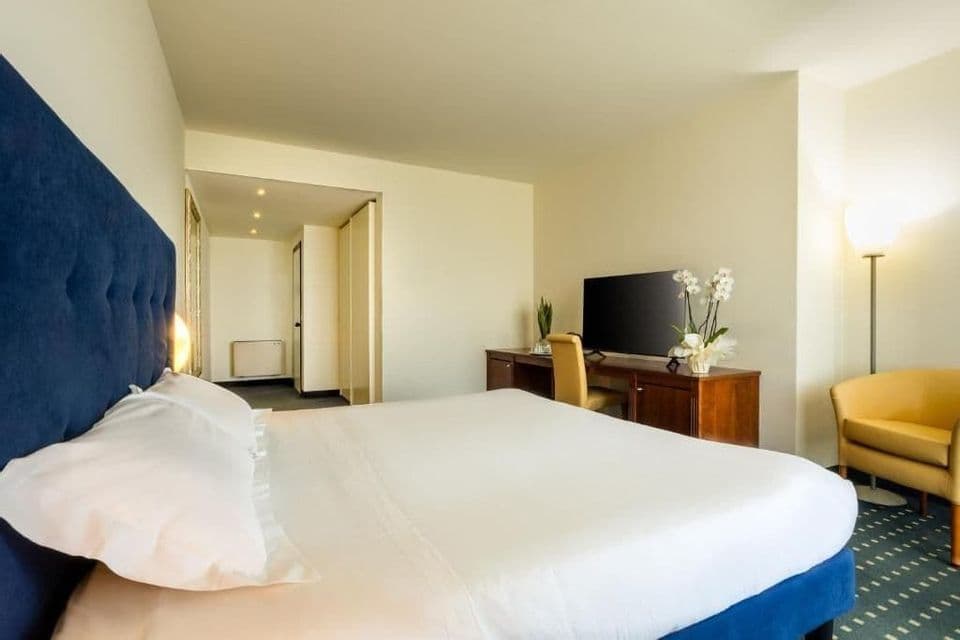 Hotel room with a large bed, blue headboard, wooden desk with a TV, and a yellow armchair in the corner.