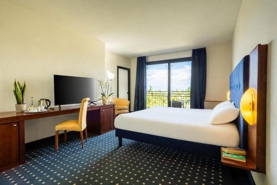 Hotel room with a large bed, wooden desk with a TV, patterned blue carpet, and sliding balcony doors.