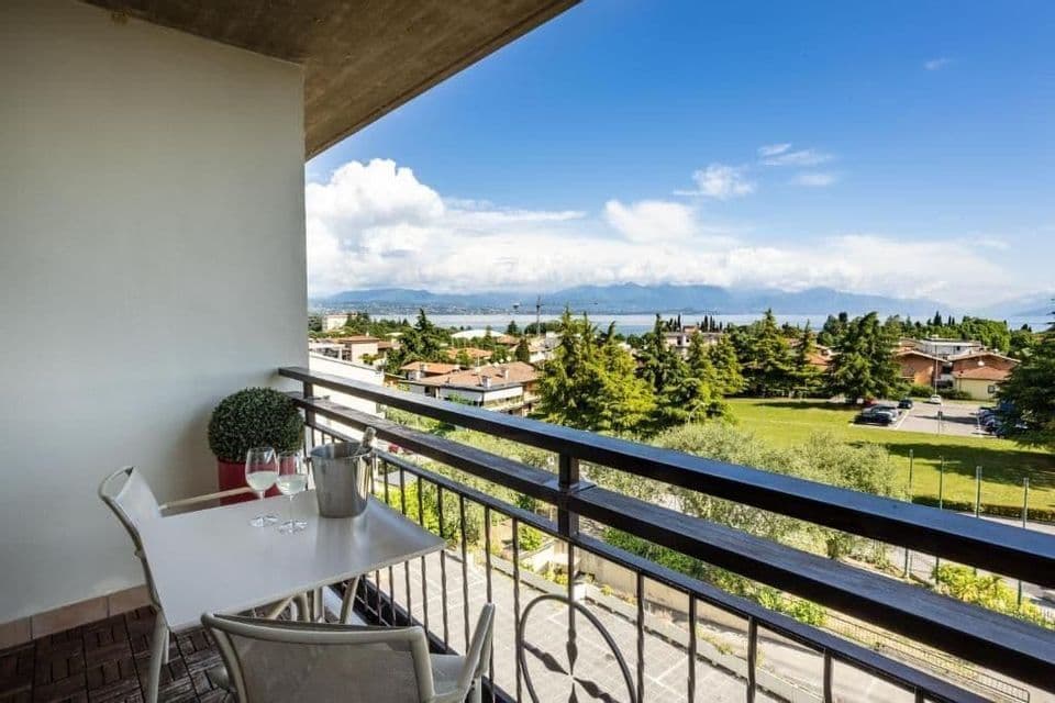 Balcony with two chairs, a small table, wine glasses, and an ice bucket overlooking green trees and a distant lake.