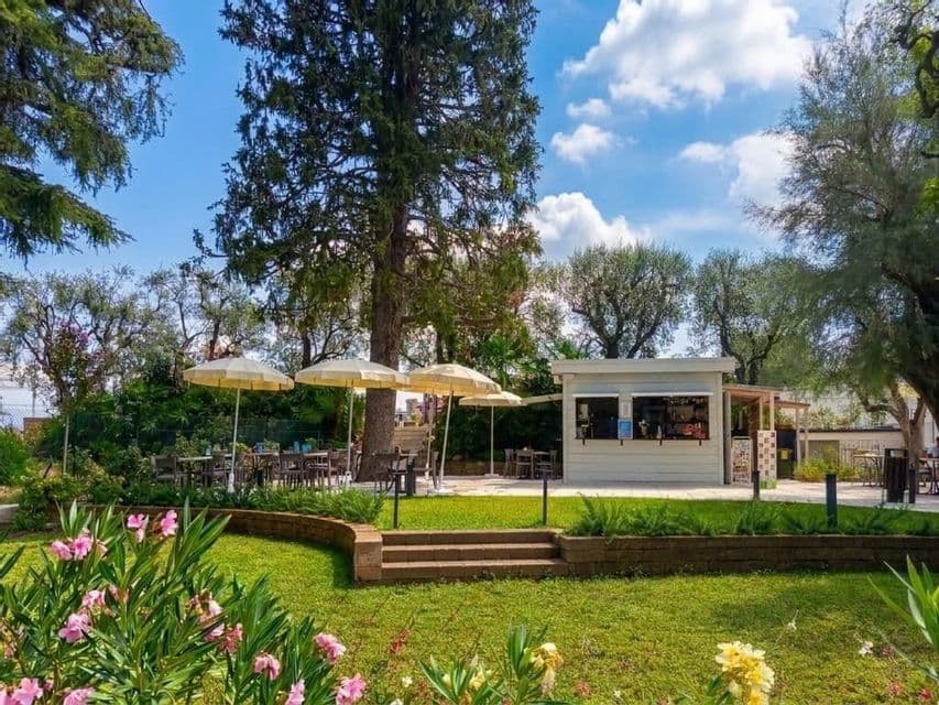 An outdoor cafe kiosk surrounded by a green lawn, tall trees, and patio seating with umbrellas under a partly cloudy sky.