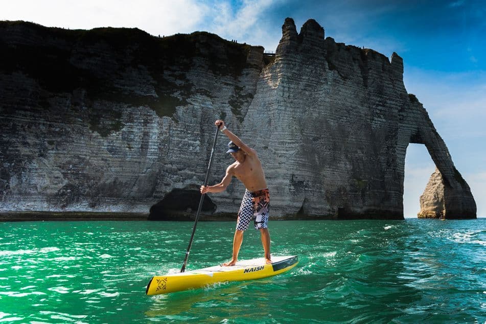 Ein Mann auf einem Stand-Up Paddleboard paddelt in türkisfarbenem Wasser neben einer großen Klippenformation mit einem natürlichen Bogen.