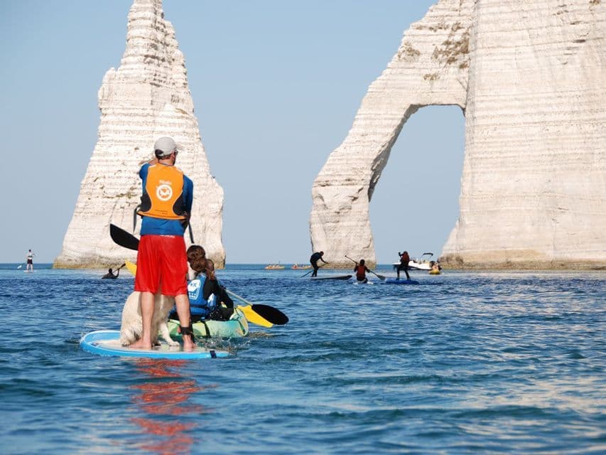 Ein Mann mit Hund auf einem Paddleboard und andere beim Kajakfahren nahe hoher, weißer Seeklippen mit einem natürlichen Bogen.