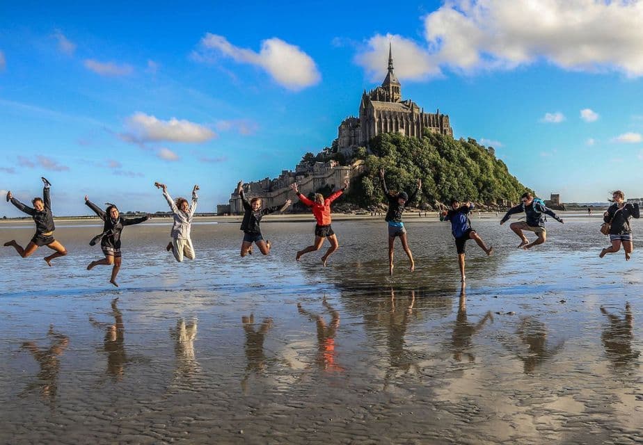 Eine WeRoad-Gruppe springt gemeinsam an einem nassen Sandstrand, ihre Spiegelungen im Wasser sichtbar und eine Burg auf einem Hügel im Hintergrund.