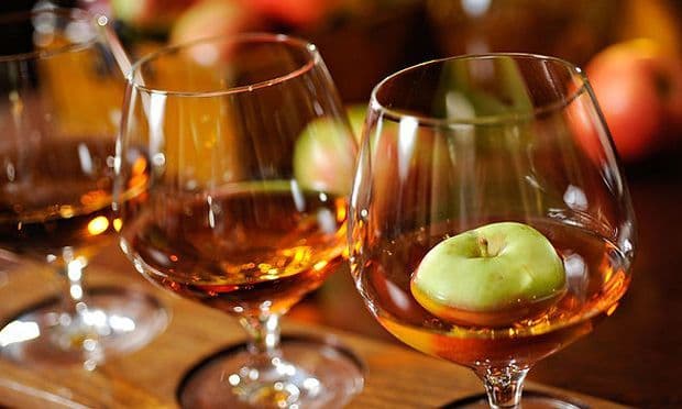 Nahaufnahme eines kleinen grünen Apfels, der in einem mit bernsteinfarbener Flüssigkeit gefüllten Weinbrandglas schwimmt, daneben weitere Gläser für eine Verkostung aufgereiht.