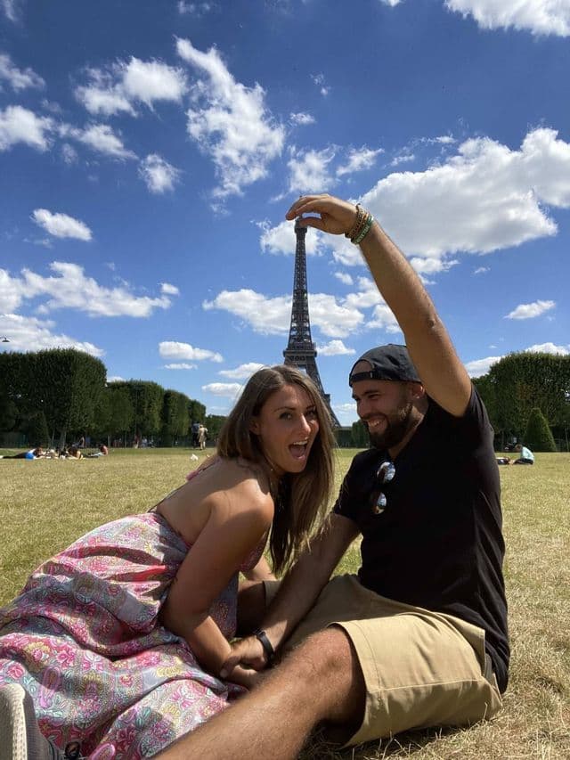 Ein Paar sitzt auf einer grünen Wiese, wobei der Mann spielerisch posiert, als würde er den Eiffelturm vor blauem Himmel halten.