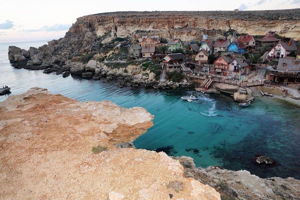 Vista dall'alto di un villaggio colorato con case rustiche incastonate in una cala rocciosa a picco sul mare, con acqua turchese e cristallina.