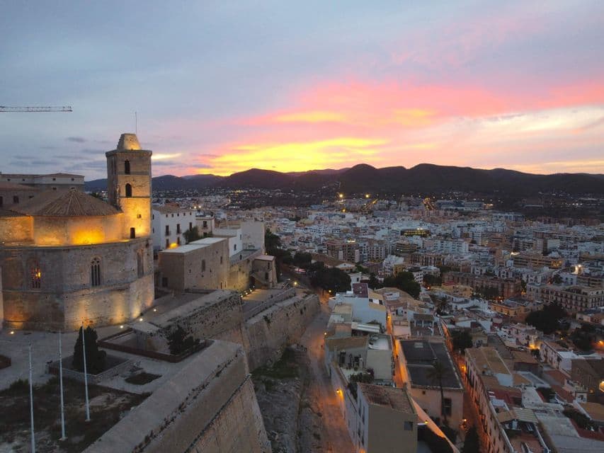 Una cattedrale in pietra illuminata domina una vasta città al tramonto, con montagne distanti sotto un cielo colorato.