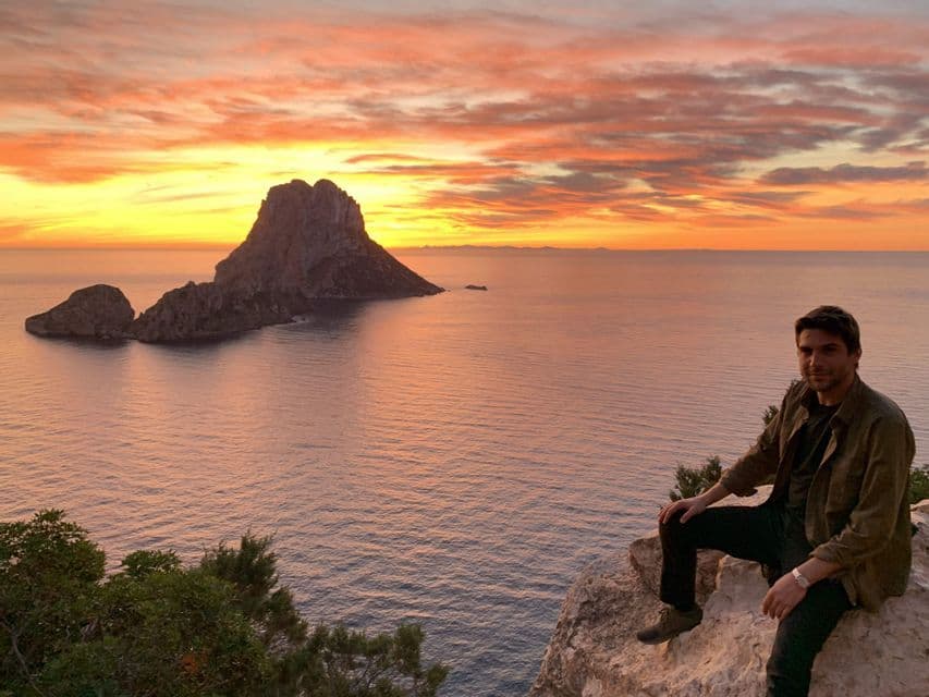 Un uomo siede su una scogliera rocciosa che si affaccia sul mare e su una grande formazione rocciosa durante un tramonto colorato.