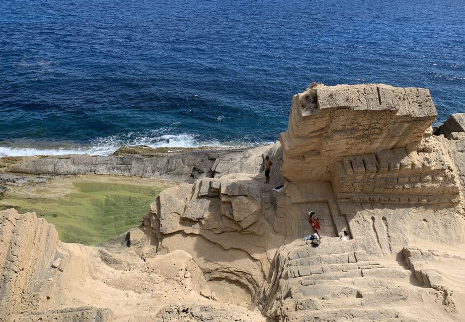 Due persone esplorano grandi formazioni rocciose sabbiose su una costa, accanto all'oceano blu profondo e a una piscina naturale verde.