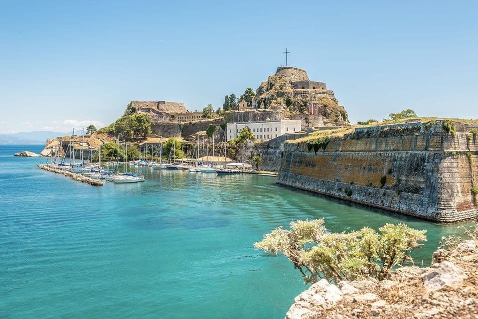 A stone fortress on a hill overlooks a marina with sailboats docked in a bay with turquoise water on a sunny day.