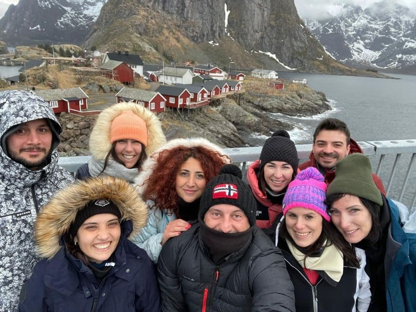 Un gruppo WeRoad in abiti invernali sorride per un selfie con un villaggio costiero e montagne innevate sullo sfondo.