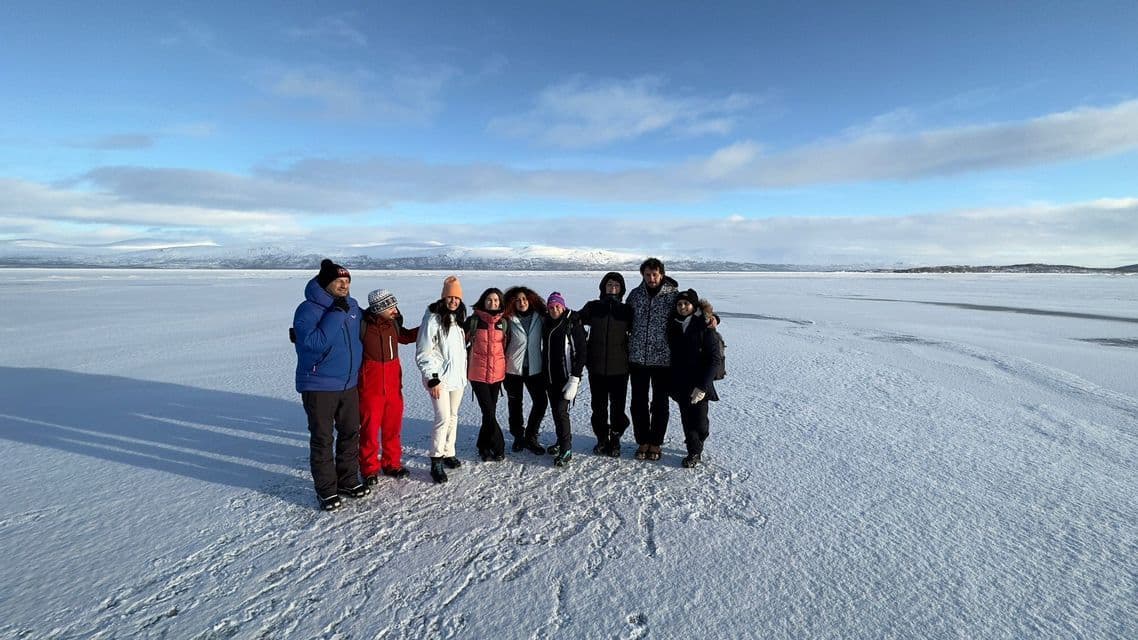 Un gruppo WeRoad in viaggio posa insieme su una vasta pianura innevata, con montagne distanti sotto un cielo blu.
