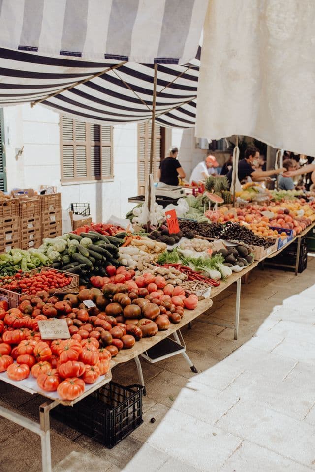 Una variedad de verduras y frutas frescas se exhiben en una mesa larga en un puesto de mercado al aire libre bajo un toldo a rayas.